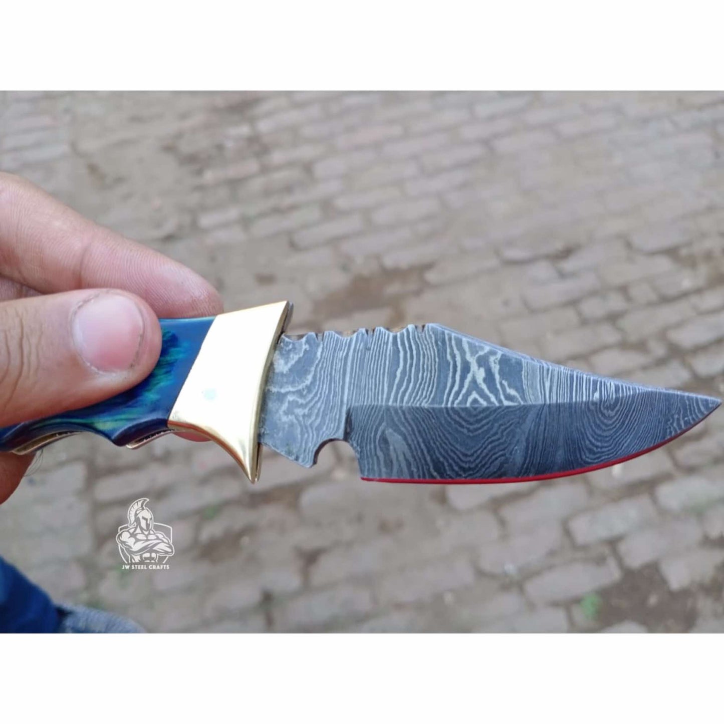 Close-up side view of an open Damascus steel folding knife blade held in hand, showing layered Damascus patterns, a sharp drop-point profile with jimping near the spine, a red-accented cutting edge, and a polished brass bolster transitioning into a blue color-wood handle, photographed outdoors with a blurred brick ground background and JW SteelCrafts watermark.