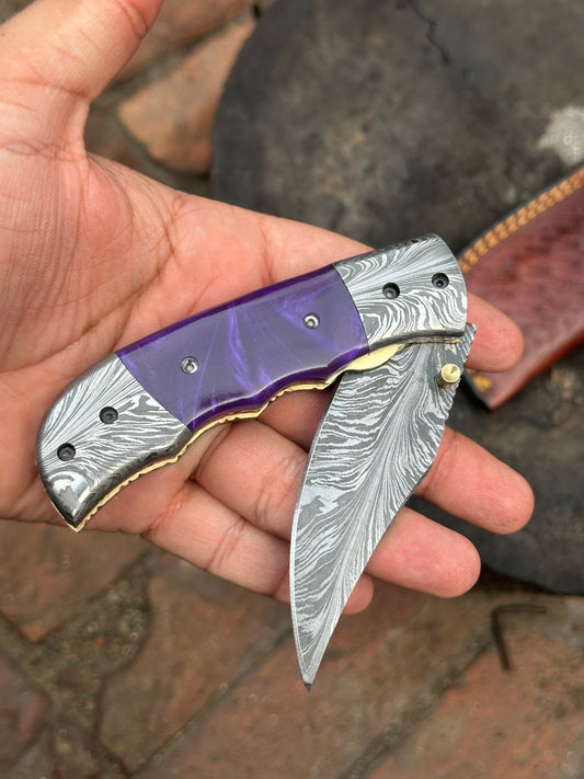 Folding knife with purple and silver handle held in a hand against a blurred background