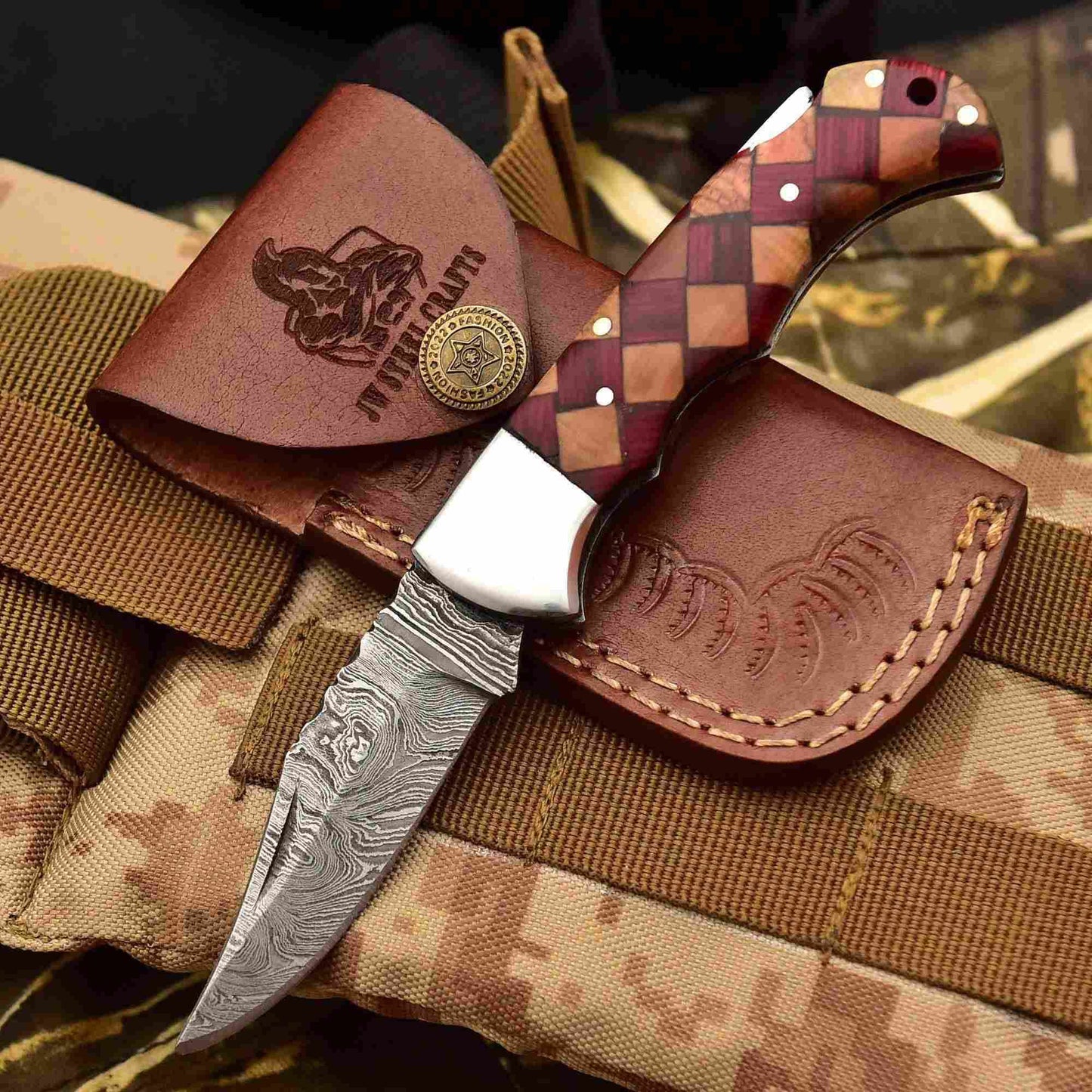 Handcrafted Damascus steel folding knife with checkered red and natural wood handle, displayed with a brown leather sheath on tactical fabric background by JW SteelCrafts