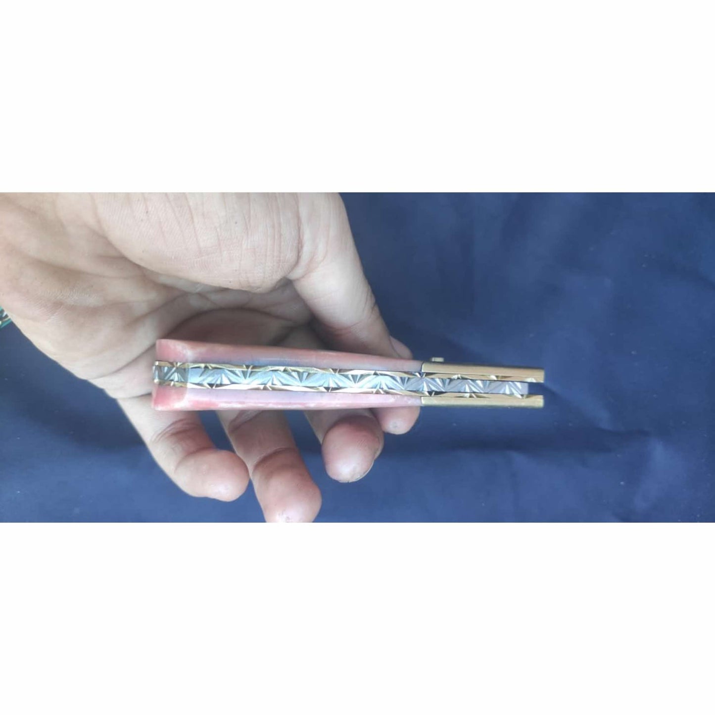 Side profile of a handmade Damascus steel folding knife held in hand, showing the closed position with pink stone-style handle scales, decorative patterned spine filework, brass liners, and a dark blue fabric background.