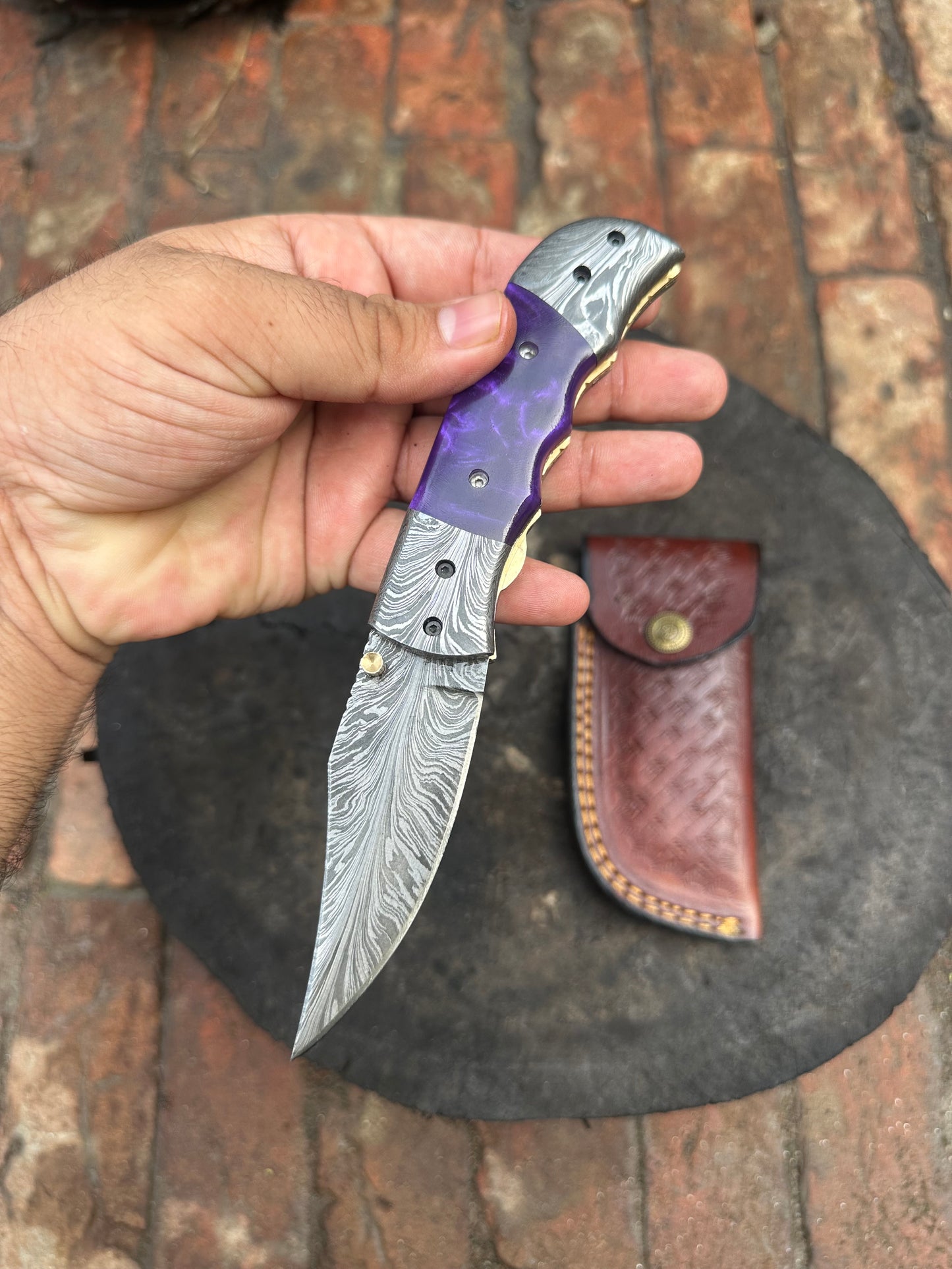 Hand holding a Damascus steel knife with a purple handle, leather sheath in the background.
