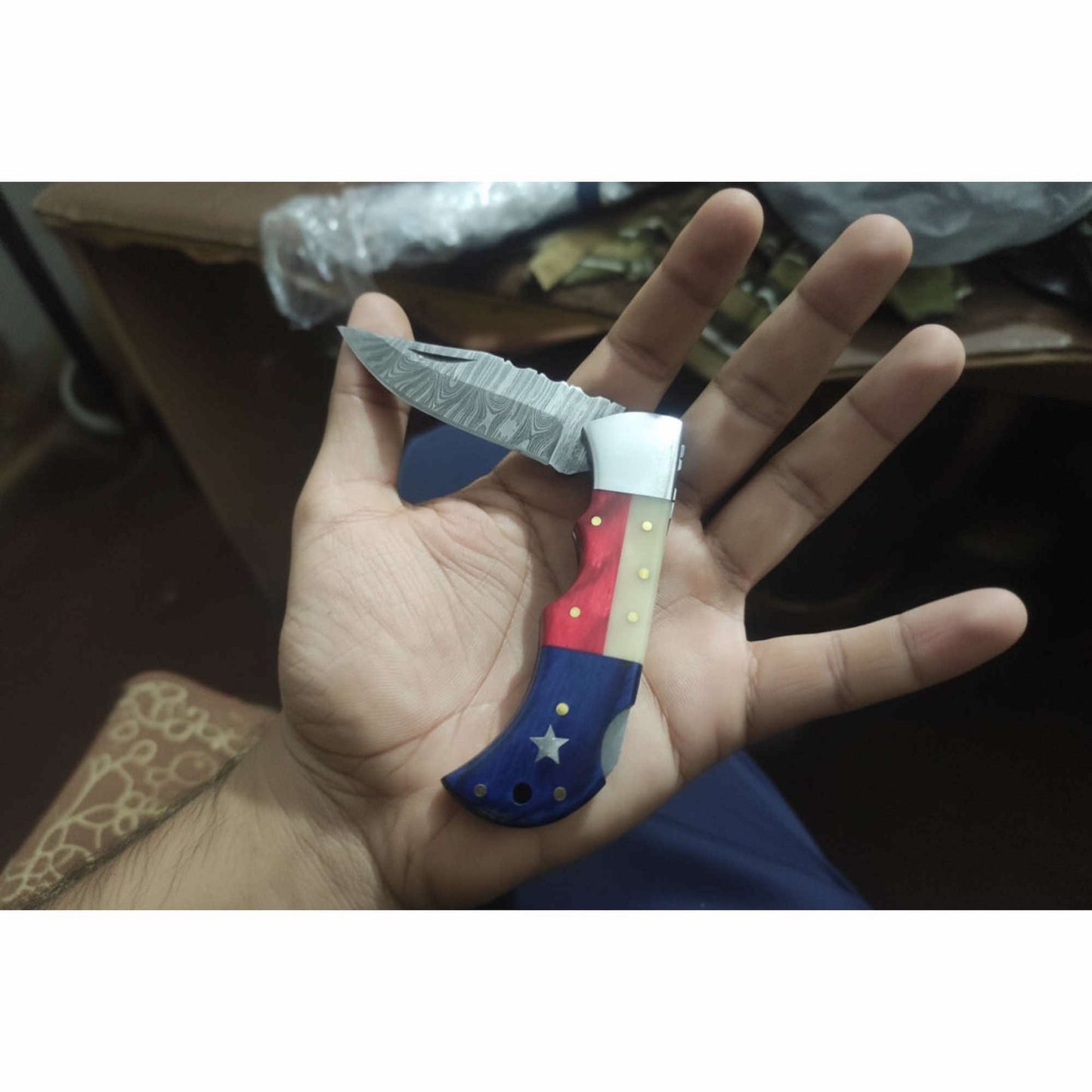 Custom Damascus steel folding knife with patriotic red white and blue handle, star inlay grip and etched blade displayed in hand during finishing stage