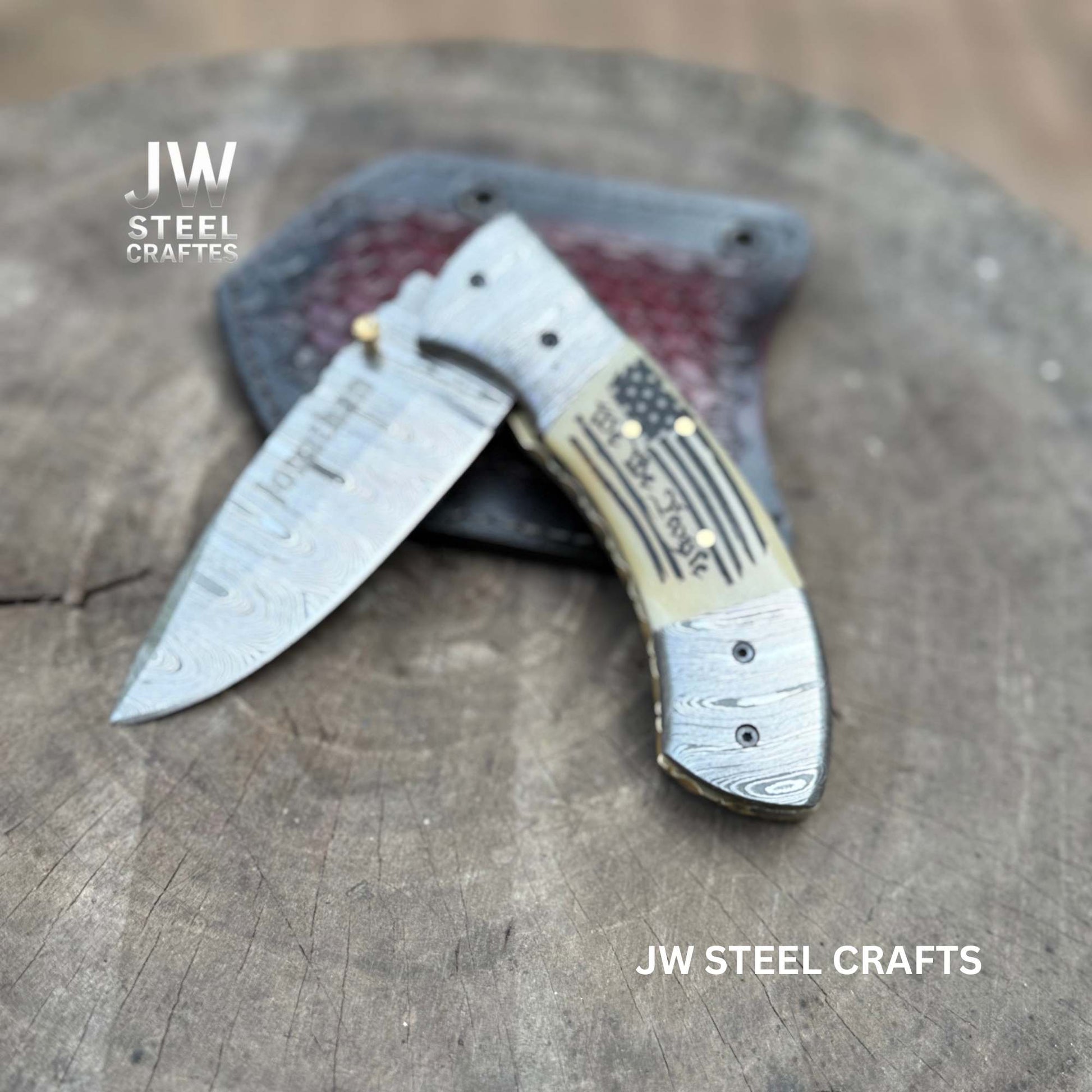 Folding knife with American flag handle on a wooden surface, branded 'JW Steel Crafts'.