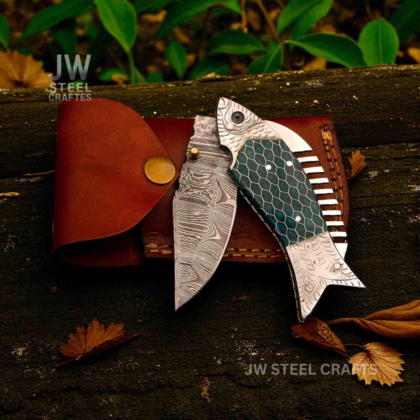 Fish-shaped Damascus steel folding knife with green honeycomb handle, shown partially open beside a brown leather sheath on a rustic wooden surface, handcrafted by JW SteelCrafts.