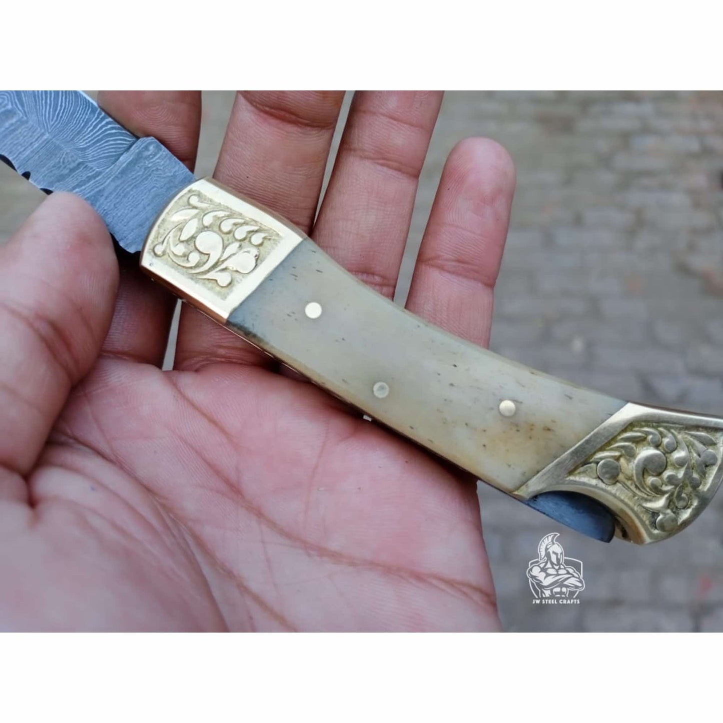 Close-up of a handcrafted Damascus steel folding knife handle featuring a smooth light bone grip with brass pins and engraved brass bolsters, shown resting in a hand with the blade partially visible, photographed outdoors with a subtle brick background and JW SteelCrafts watermark.