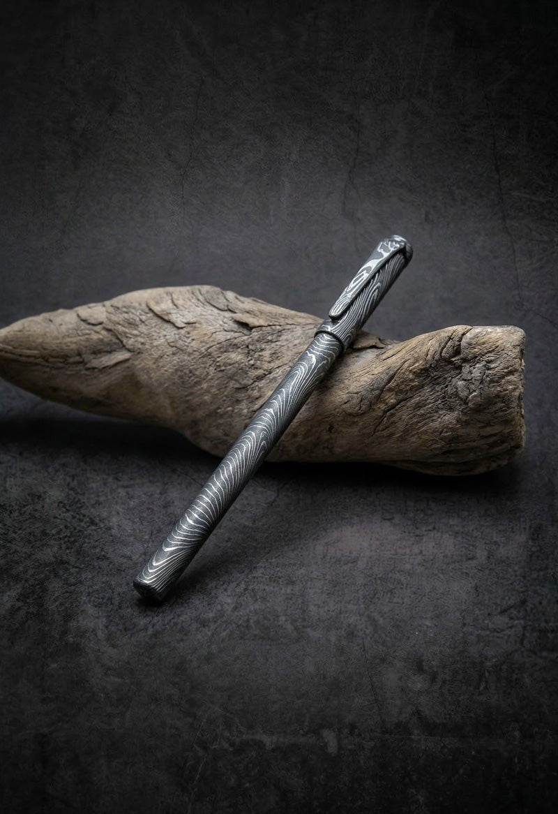 Handmade Damascus steel twist pattern pen resting on driftwood – JW Steel Crafts custom artisan Damascus steel pen gift collection