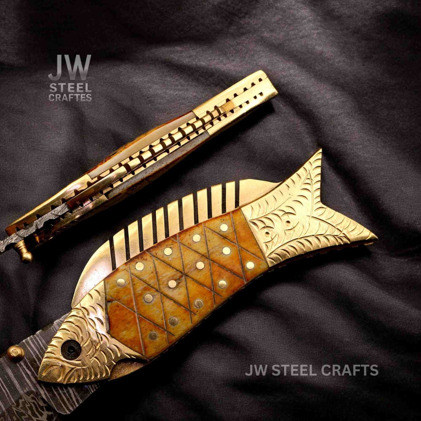 JW SteelCrafts Damascus steel fish folding knife with engraved brass handle and natural bone inlay, showing spine jimping and detailed craftsmanship on a dark fabric background.