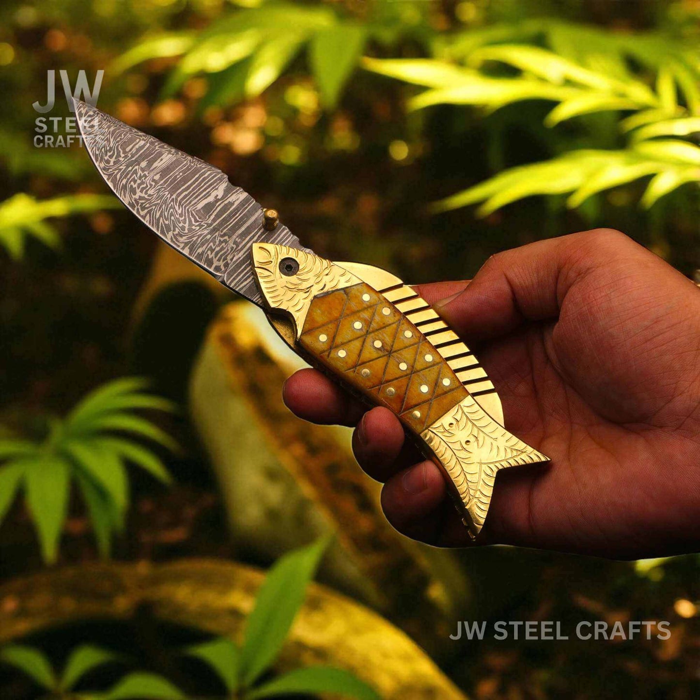 JW SteelCrafts handmade fish-shaped Damascus steel folding knife with engraved brass handle and natural bone inlay, shown held in hand outdoors.