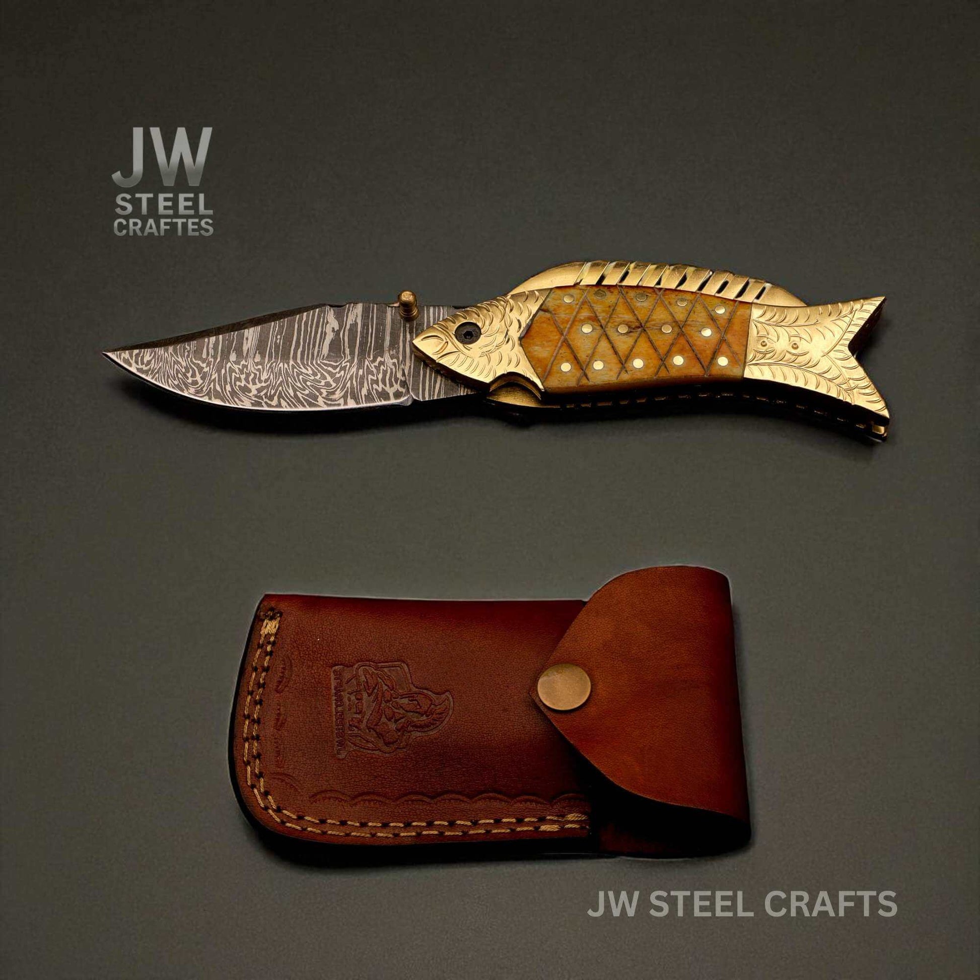 JW SteelCrafts Damascus steel fish folding knife with engraved brass handle and natural bone inlay, shown open above a handmade brown leather sheath on a dark background.