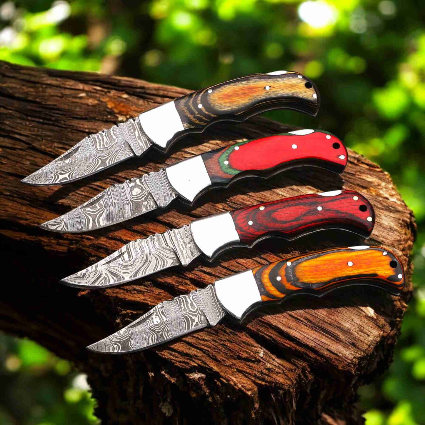Four hand-forged Damascus steel folding pocket knives by JW SteelCrafts with multicolor pakka wood handles (brown, red, dark red, and orange), steel bolsters, and patterned Damascus blades displayed on a rustic tree stump in outdoor forest setting