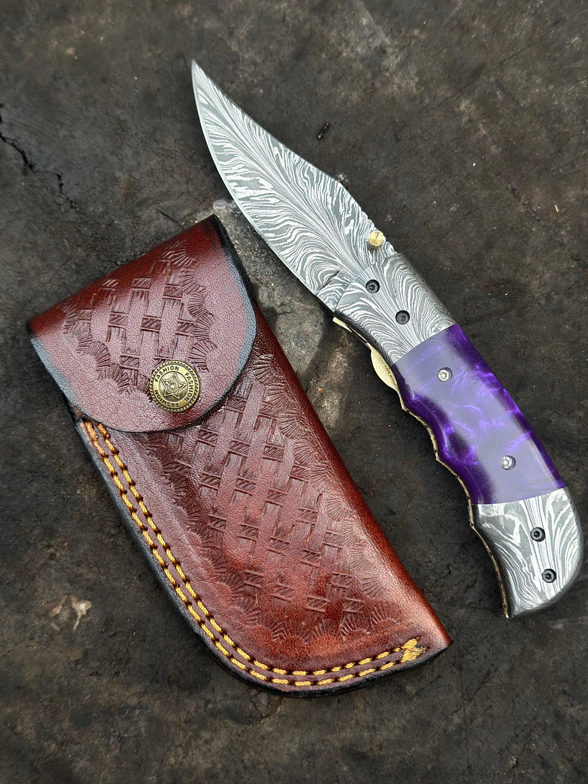 Folding knife with purple and silver handle on a brown leather sheath against a dark stone background