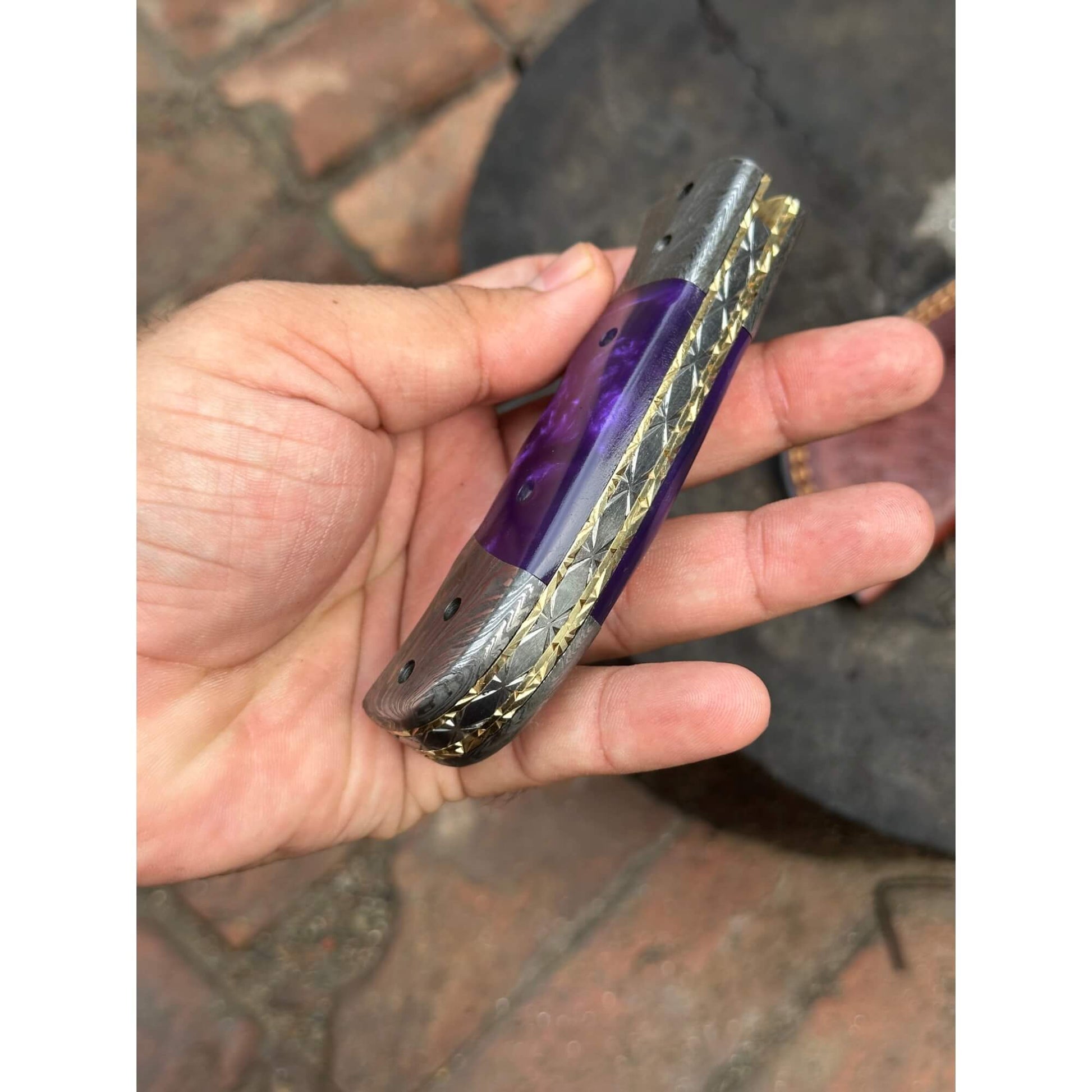 Closed Damascus steel folding knife with purple resin handle, decorative brass liner, and feather pattern steel bolsters, shown in hand – JW SteelCrafts