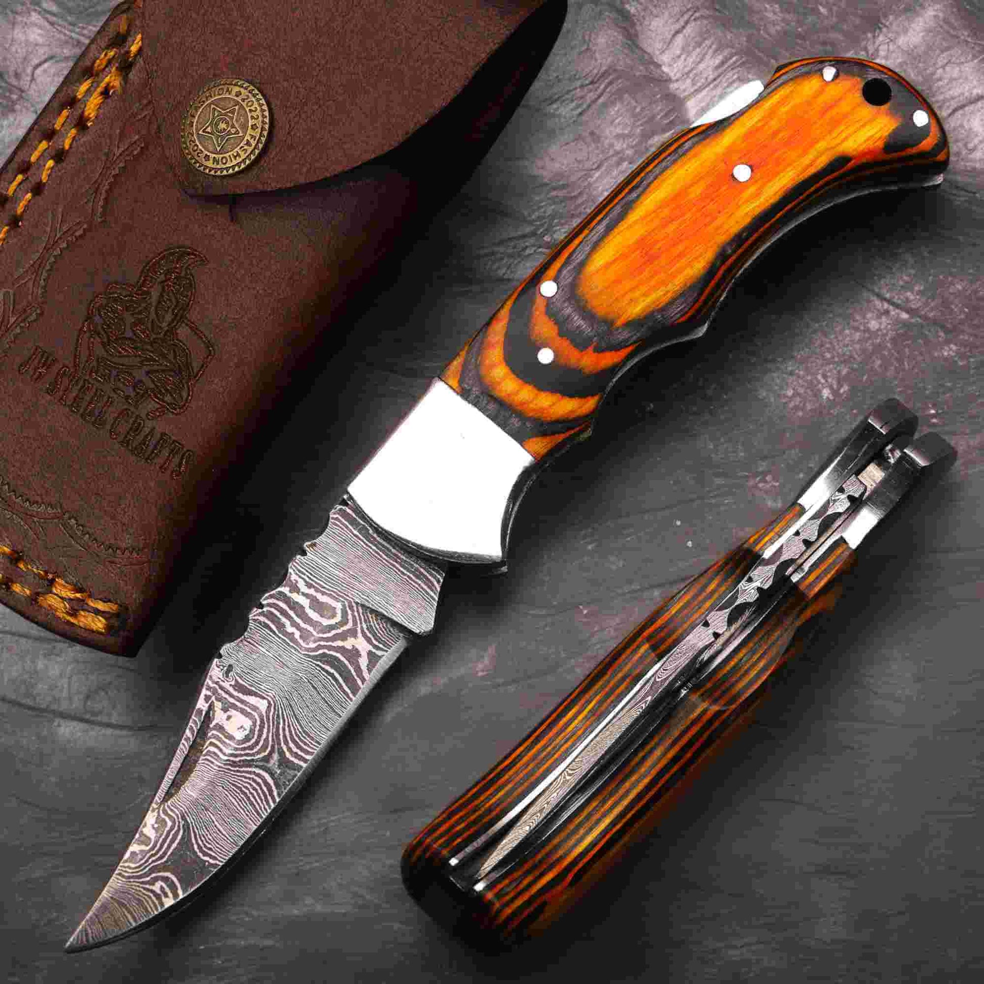 Hand-forged Damascus steel folding pocket knife by JW SteelCrafts with orange pakka wood handle, steel bolster, liner lock mechanism, patterned Damascus blade, spine filework detail, and hand-stitched leather sheath displayed on dark textured background