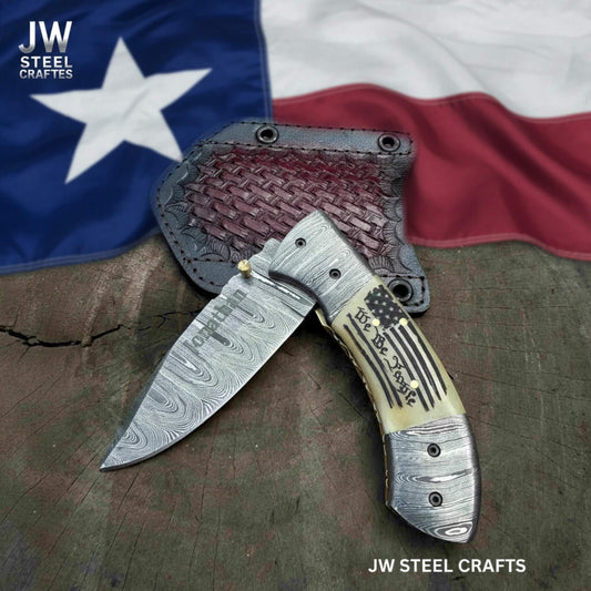 Open Damascus steel folding knife with American flag handle and engraved blade, displayed on textured leather sheath with Texas flag background by JW SteelCrafts.