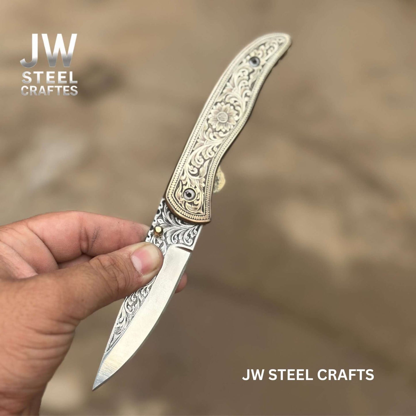 Hand-engraved brass handle folding knife by JW SteelCrafts shown partially open, featuring detailed floral engraving on the handle and hand-engraved stainless steel blade held in hand