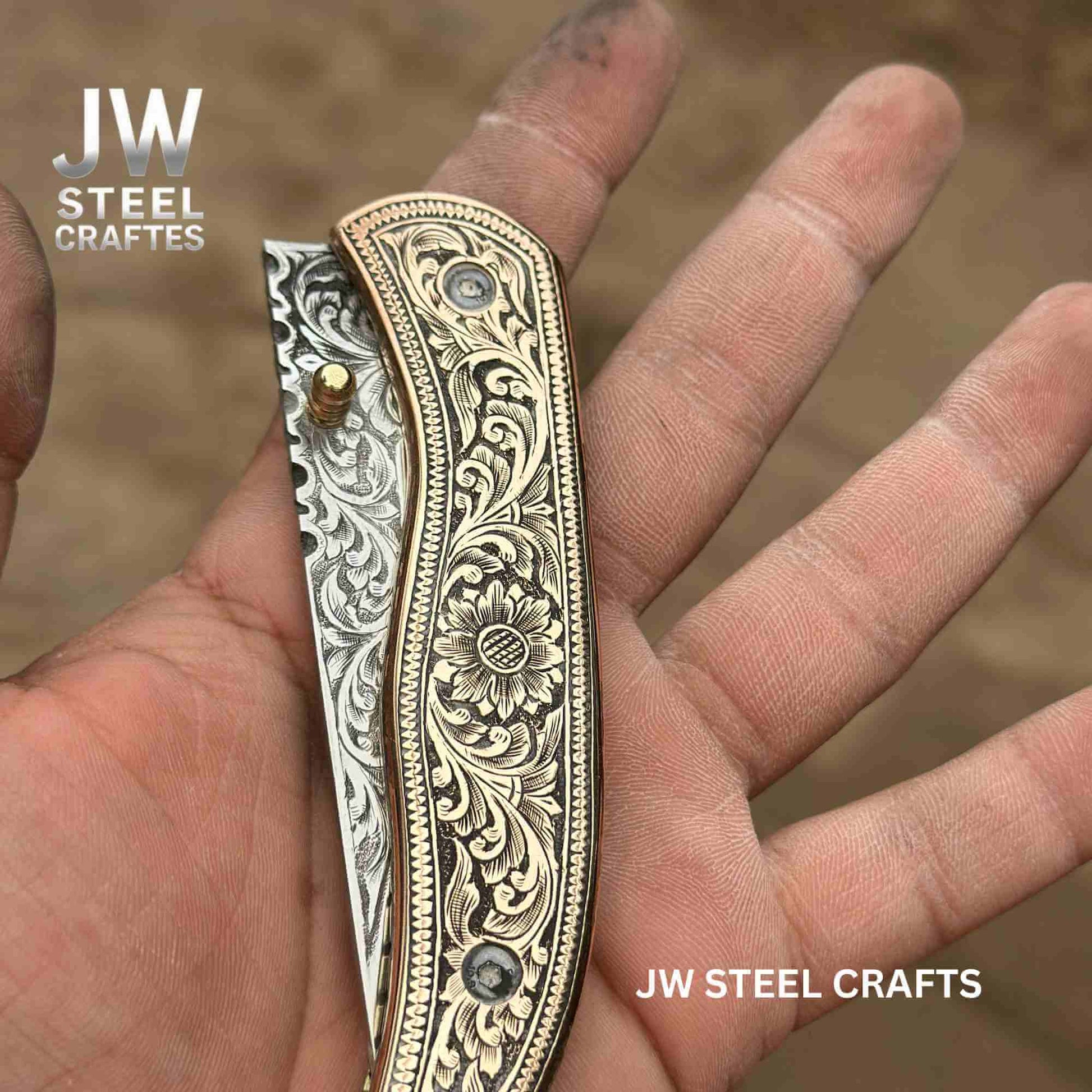 Close-up of hand-engraved brass handle folding knife by JW SteelCrafts, showcasing detailed floral engraving and partially visible engraved stainless steel blade held in hand