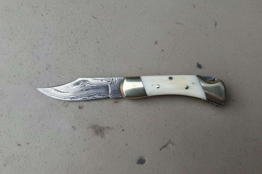 Handmade Damascus steel folding pocket knife with a curved clip-point blade showing layered Damascus patterns, polished brass bolsters, and a smooth ivory-tone handle scale, photographed on a neutral gray surface by JW SteelCrafts.