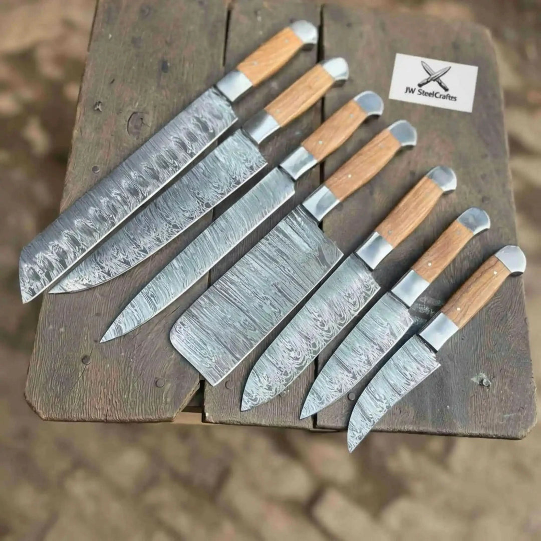 A professional 7-piece handmade Damascus steel chef knife set with light natural wood handles, including a cleaver, santoku, and utility knives, arranged on a rustic wooden surface with a JW Steel Crafts brand card.