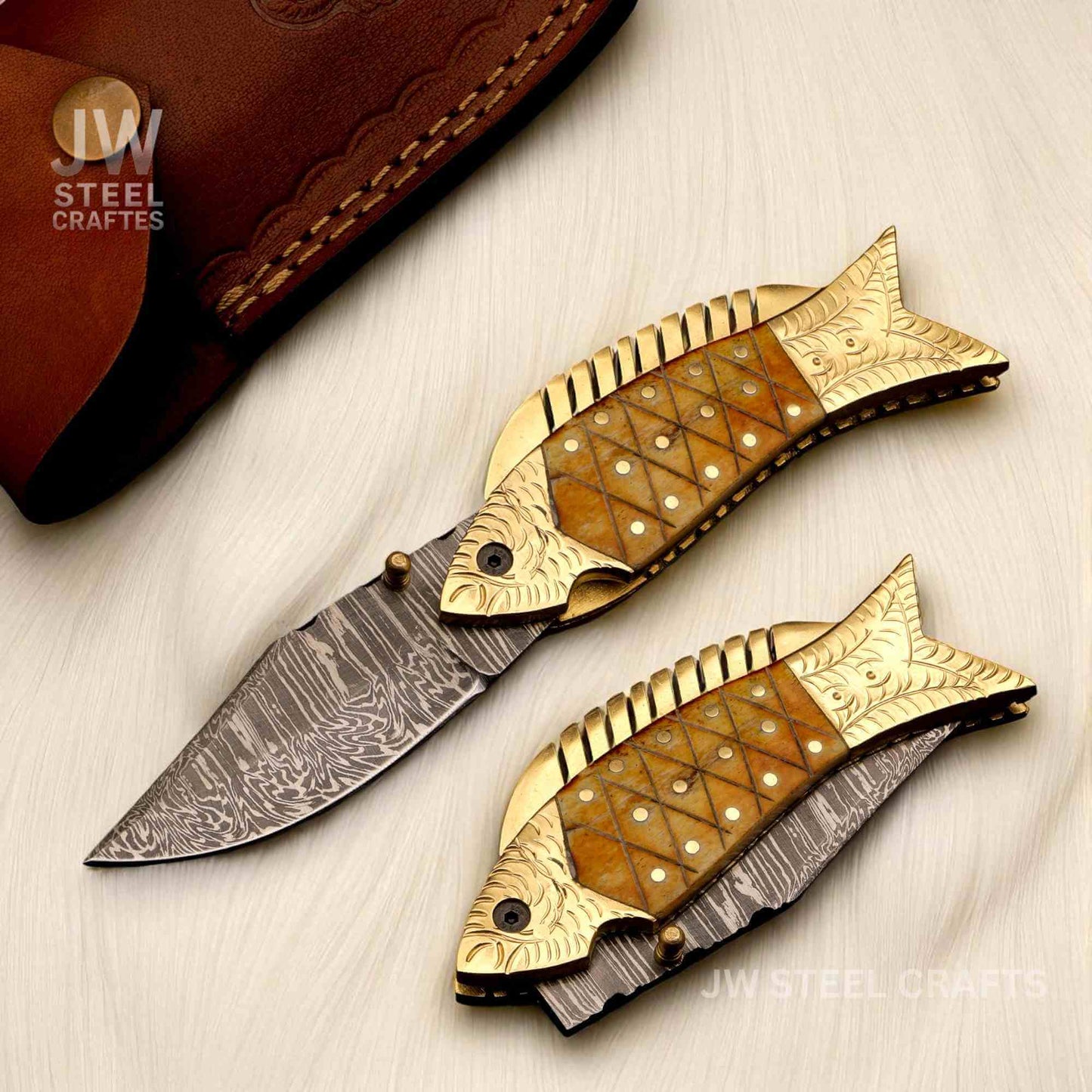 JW SteelCrafts handcrafted Damascus steel fish folding knife with brass bolster and natural bone handle, detailed engraved scales, and patterned Damascus blade displayed on a light surface.