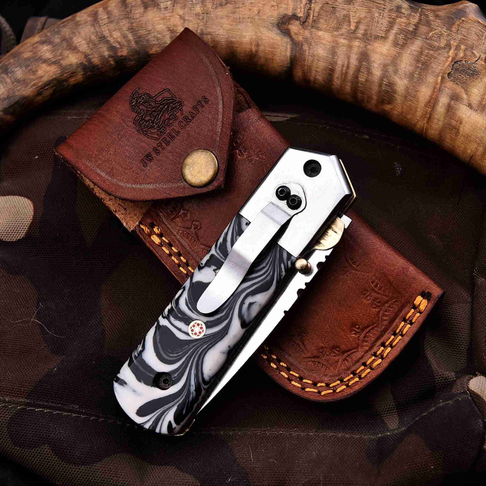 Stainless steel folding knife with marble resin handle, pocket clip, liner lock, and JW SteelCrafts leather sheath displayed on outdoor gear