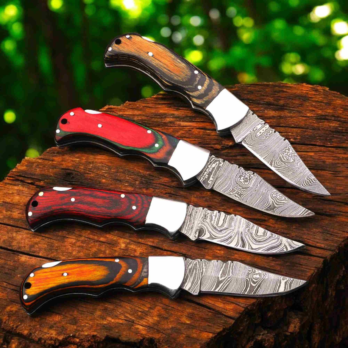 et of four hand-forged Damascus steel folding pocket knives by JW SteelCrafts featuring multicolor pakka wood handles (brown, red, dark red, and orange), steel bolsters, liner lock construction, and patterned Damascus blades displayed on a rustic wooden log in forest setting