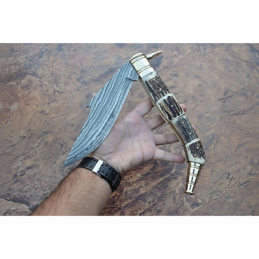 Handcrafted Damascus steel folding knife with a wide patterned blade, antler-style handle scales, and polished brass bolsters, held open in a hand against a stone-textured background by JW SteelCrafts.