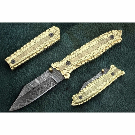 Handcrafted Damascus steel folding knife shown disassembled with engraved brass handle scales and liners, featuring ornate scrollwork detailing and a patterned Damascus blade, displayed on a dark textured fabric background by JW SteelCrafts.