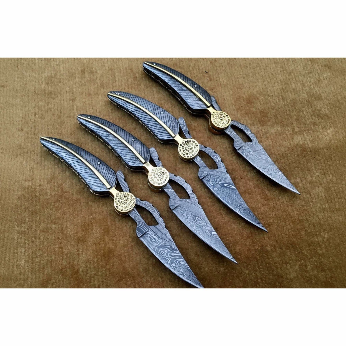 Four handmade Damascus steel folding pocket knives arranged in a row, each featuring a curved textured handle with gold accent strip, decorative round brass pivot, and patterned Damascus blade, displayed on a neutral brown background by JW SteelCrafts.