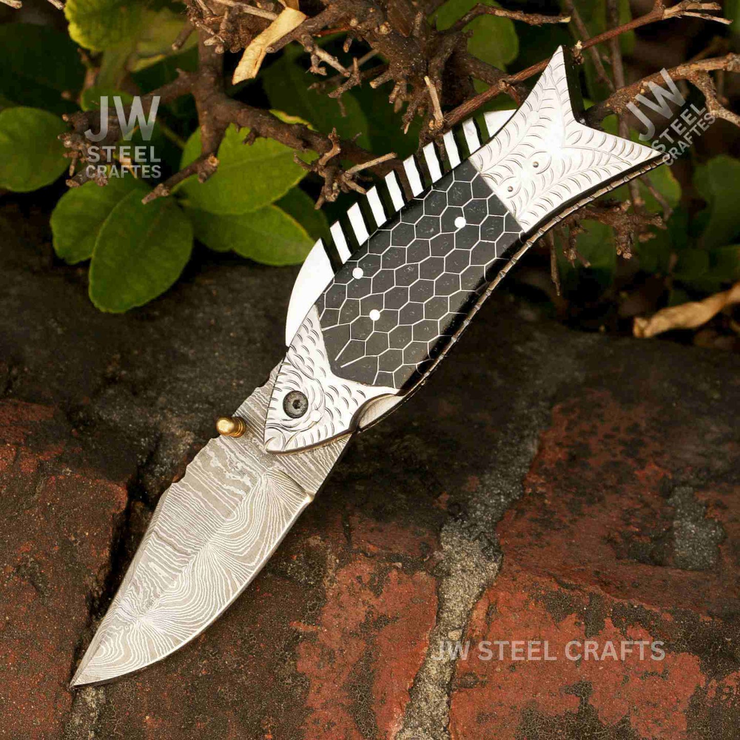 Fish-shaped Damascus steel folding knife with black honeycomb handle shown on a blacksmith forge surface with fire in the background, handcrafted by JW SteelCrafts.