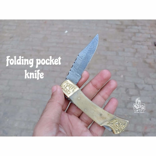 Open Damascus steel folding pocket knife held in hand, featuring a patterned Damascus blade with a slim drop-point profile, engraved brass bolsters at both ends, and a cream-colored bone-style handle scale, photographed outdoors against a brick ground background with “folding pocket knife” text and JW SteelCrafts watermark visible.