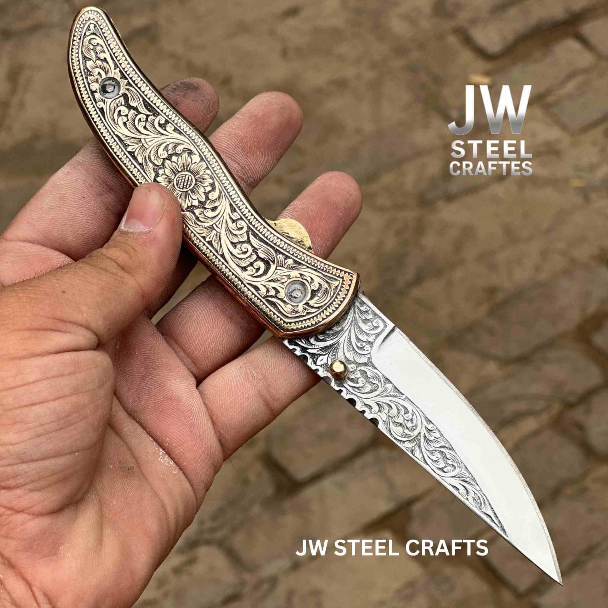 Hand-engraved folding knife by JW SteelCrafts featuring a brass handle with detailed floral engraving and a stainless steel blade, liner lock design, shown held in hand outdoors