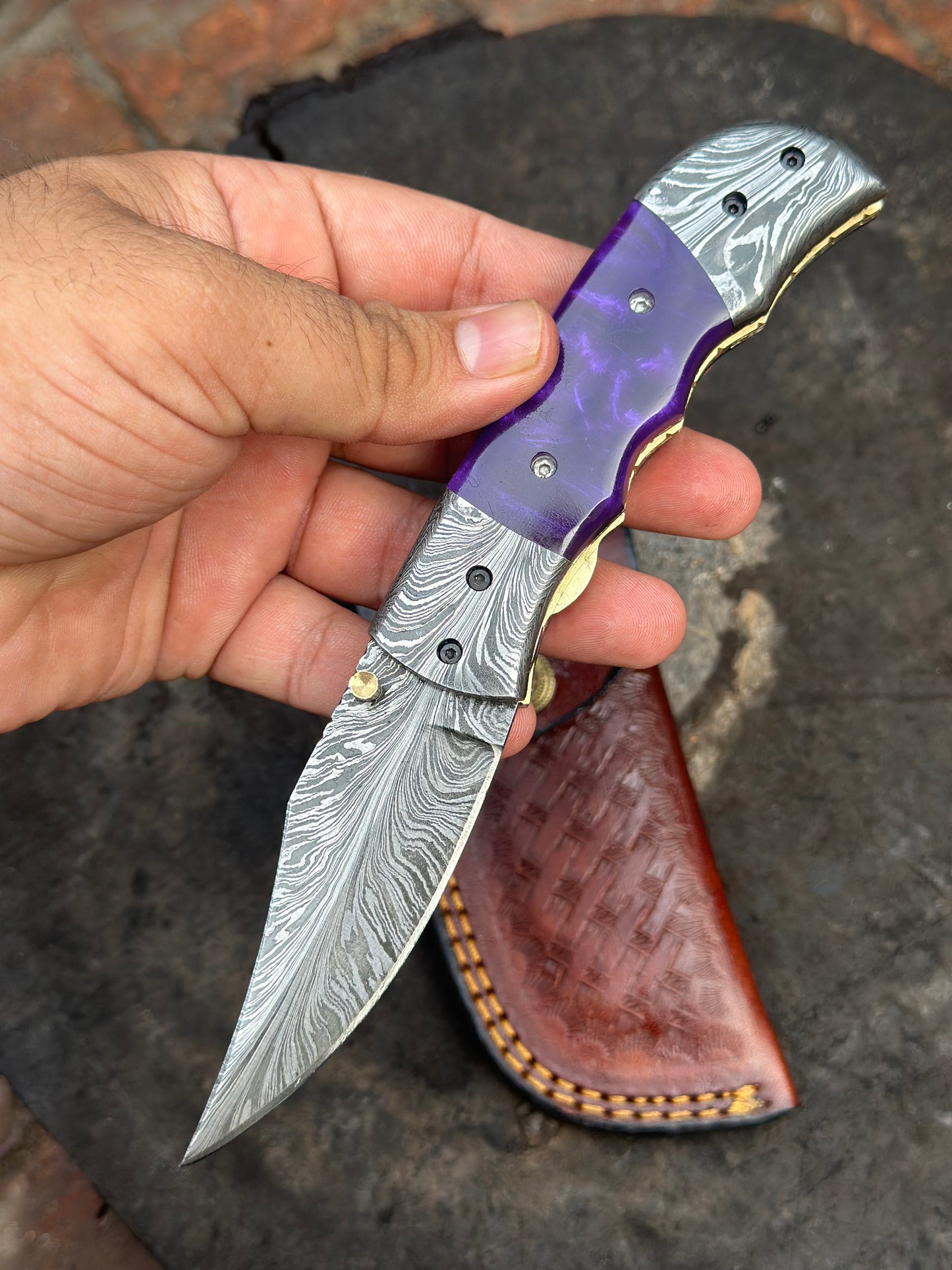 Folding knife with purple handle held by a hand, leather sheath in the background