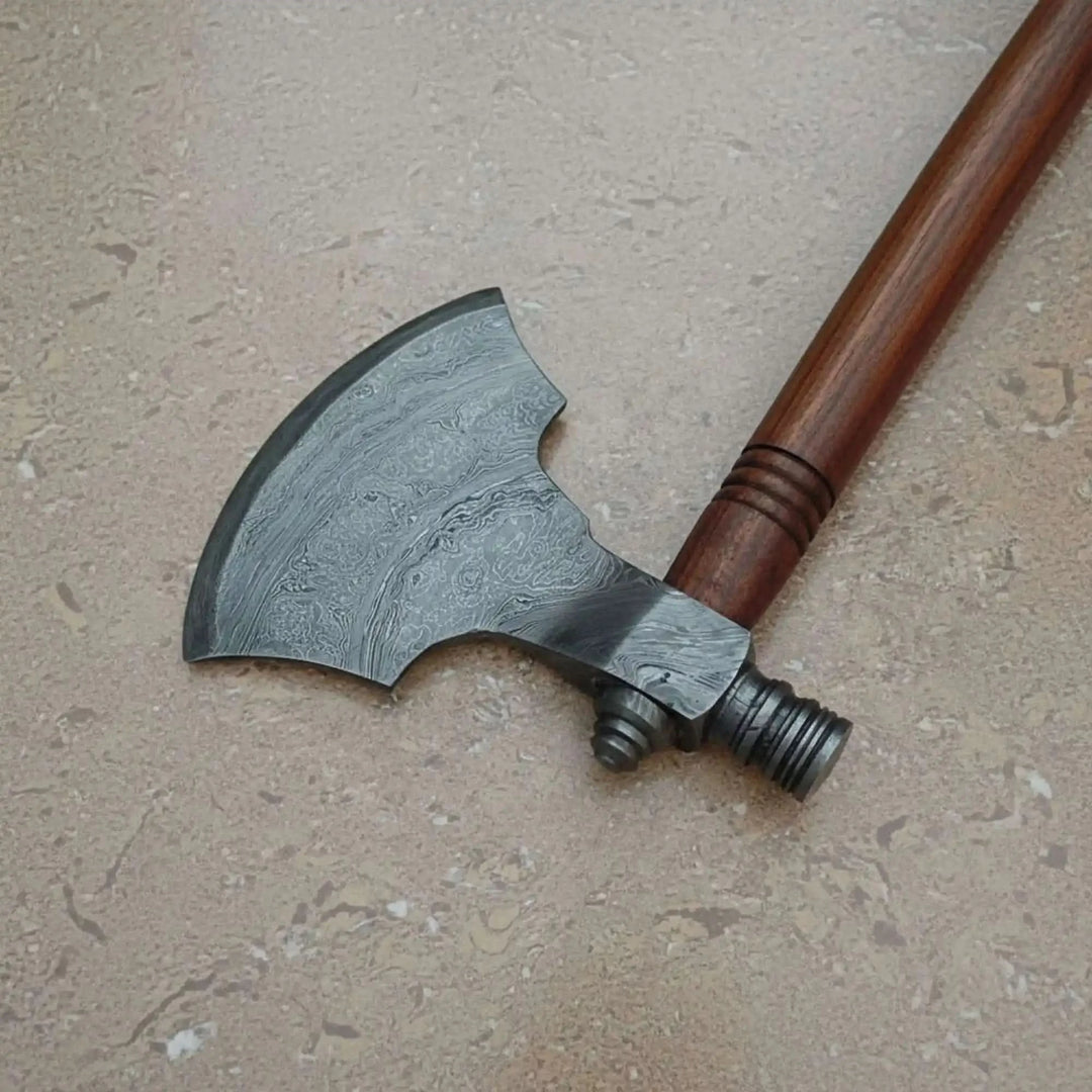     Battleforged custom Damascus hatchet with decorative layered blade and dark wooden shaft displayed on a neutral stone background
