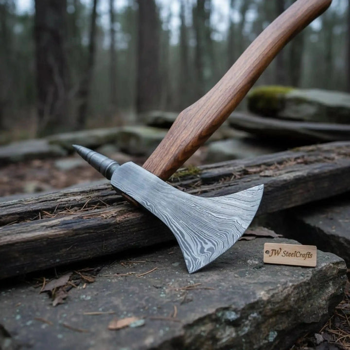     Rustic camping hatchet with Damascus steel edge and pointed rear spike resting on aged wooden logs in a misty outdoor forest setting.