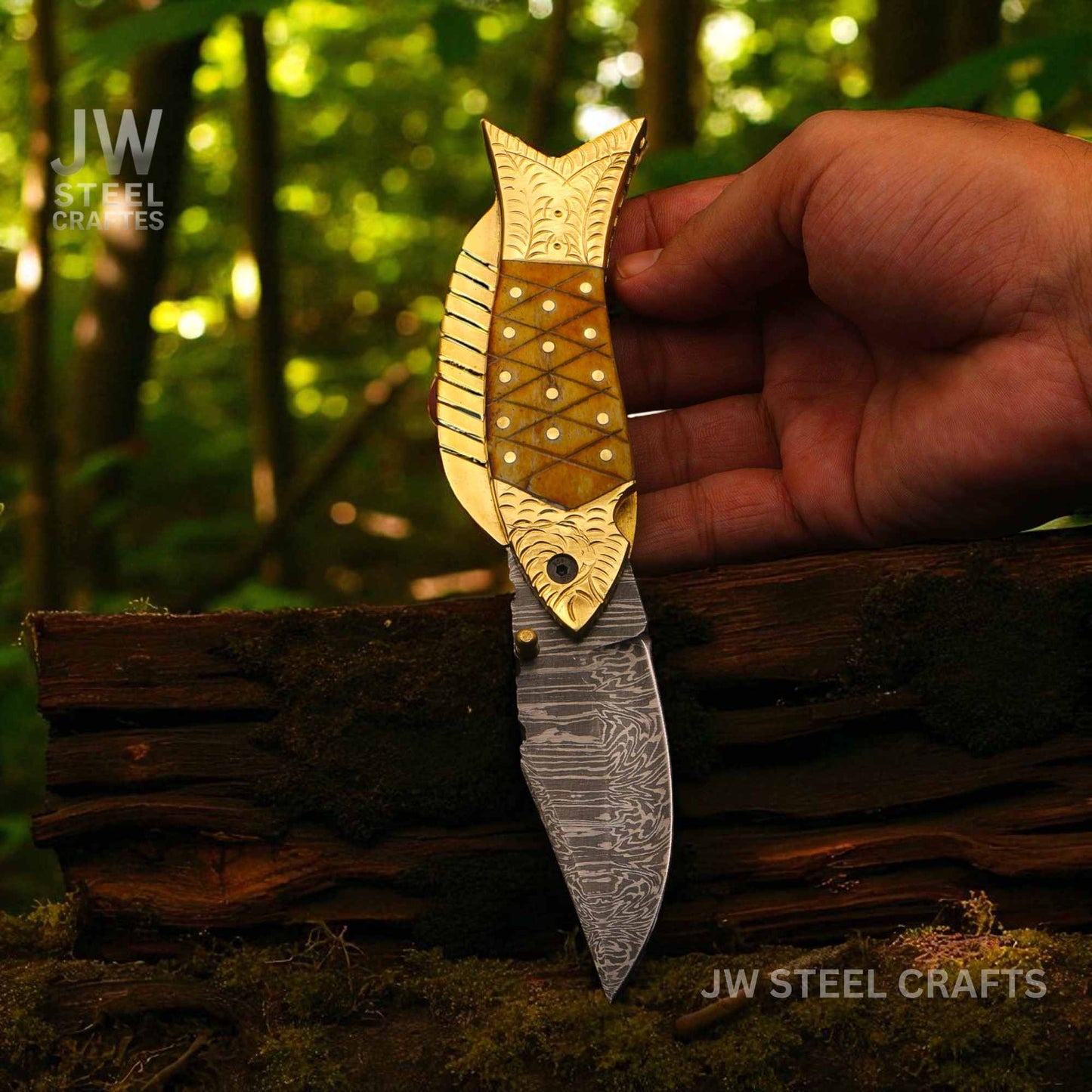 Fishing-themed knife with gold fish head held by a hand against a forest background, branded 'JW Steel Crafts'.