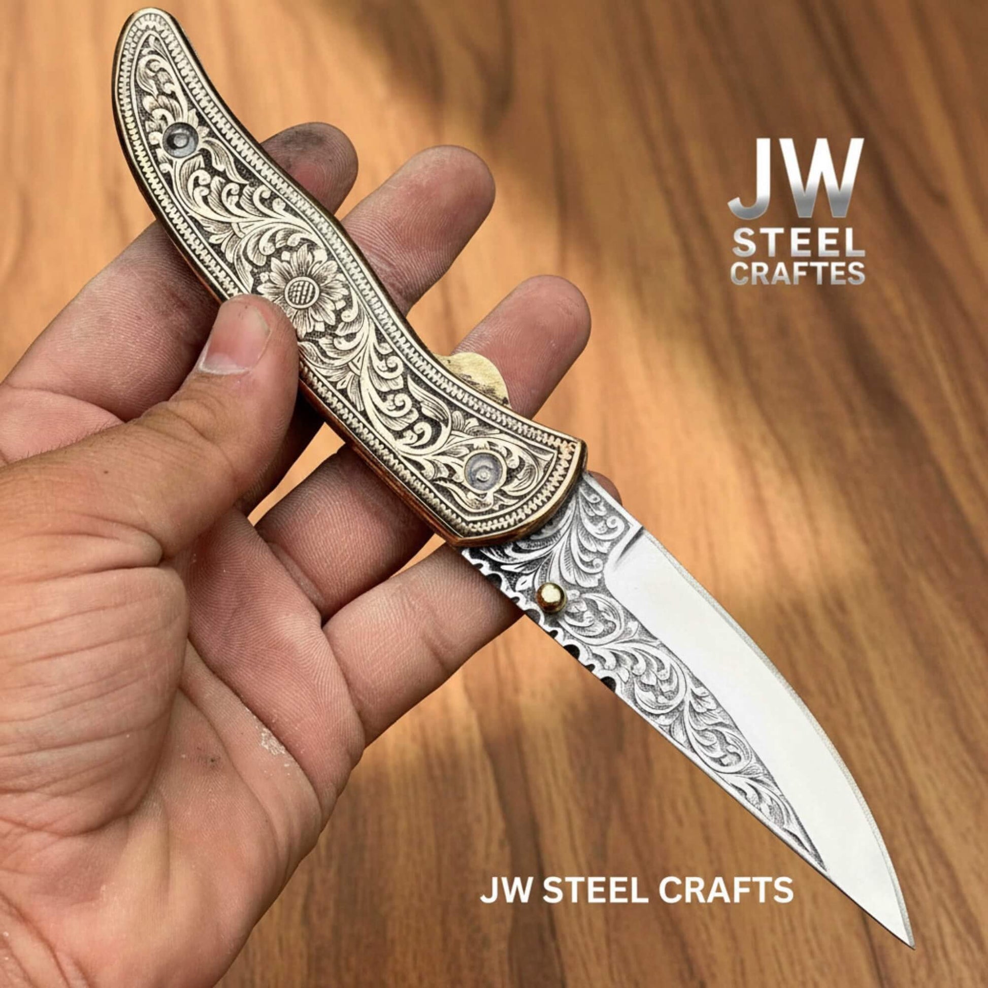 Hand-engraved brass handle folding knife by JW SteelCrafts shown in hand on a wooden surface, featuring detailed floral engraving on the brass scales, a hand-engraved stainless steel blade, brass thumb stud, and secure liner lock mechanism