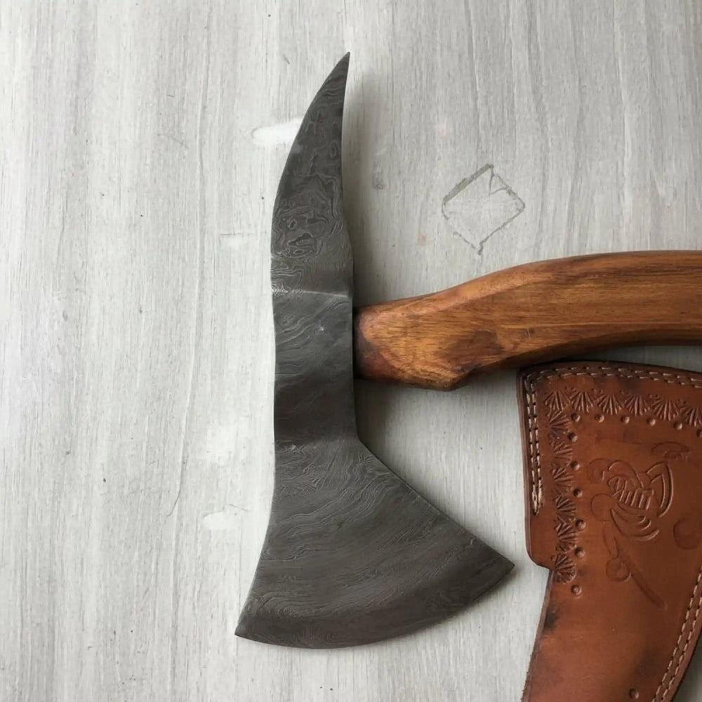     Cinderfang artisan survival hatchet featuring patterned Damascus blade and curved hardwood grip near evening flames.