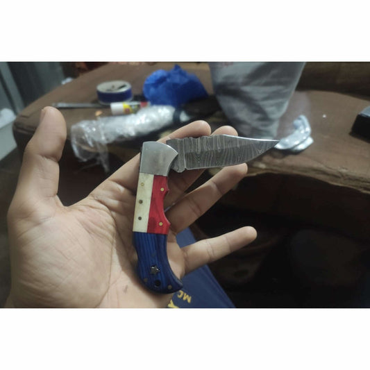 Handcrafted Damascus steel folding knife with tricolor handle in red white and blue, compact curved blade shown resting in an artisan workshop hand