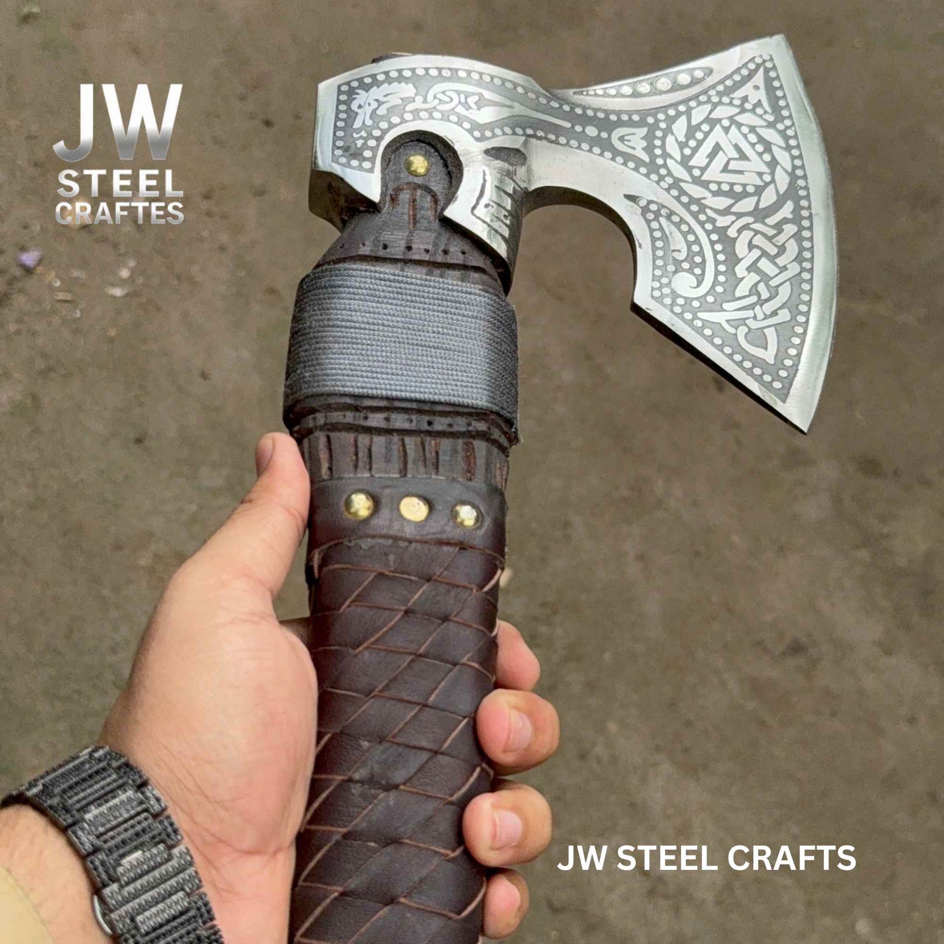 Hand holding a decorative axe with a braided leather handle against a neutral background, branded 'JW Steel Crafts'.