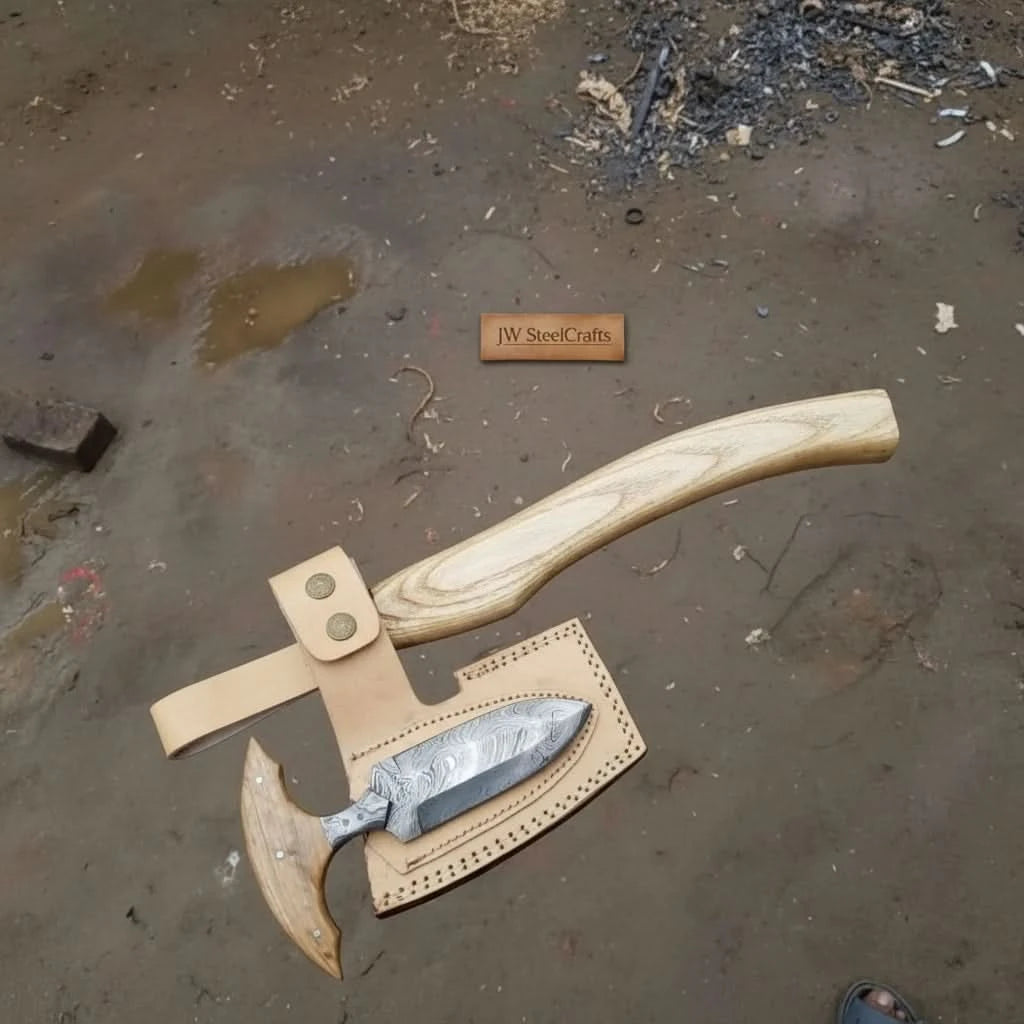     Driftstone forged Damascus steel woodland axe with etched blade detailing, pale wood handle, and handcrafted leather sheath outdoors.