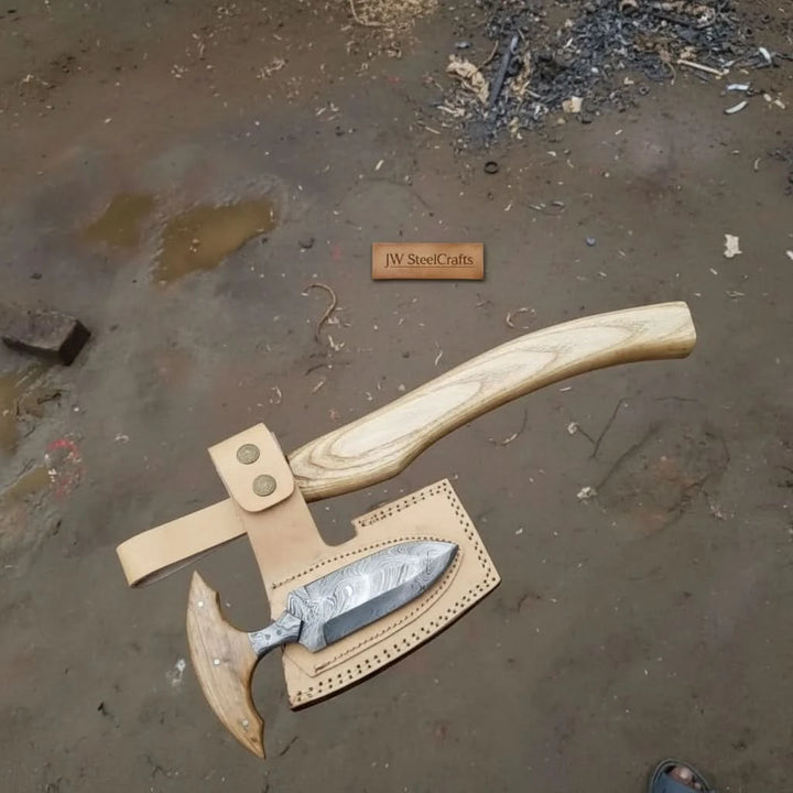     Driftstone forged Damascus steel woodland axe with etched blade detailing, pale wood handle, and handcrafted leather sheath outdoors.
