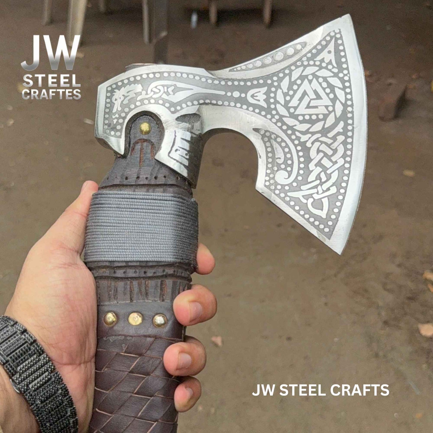 Decorative axe with intricate patterns held by a hand, branded 'JW Steel Crafts'.