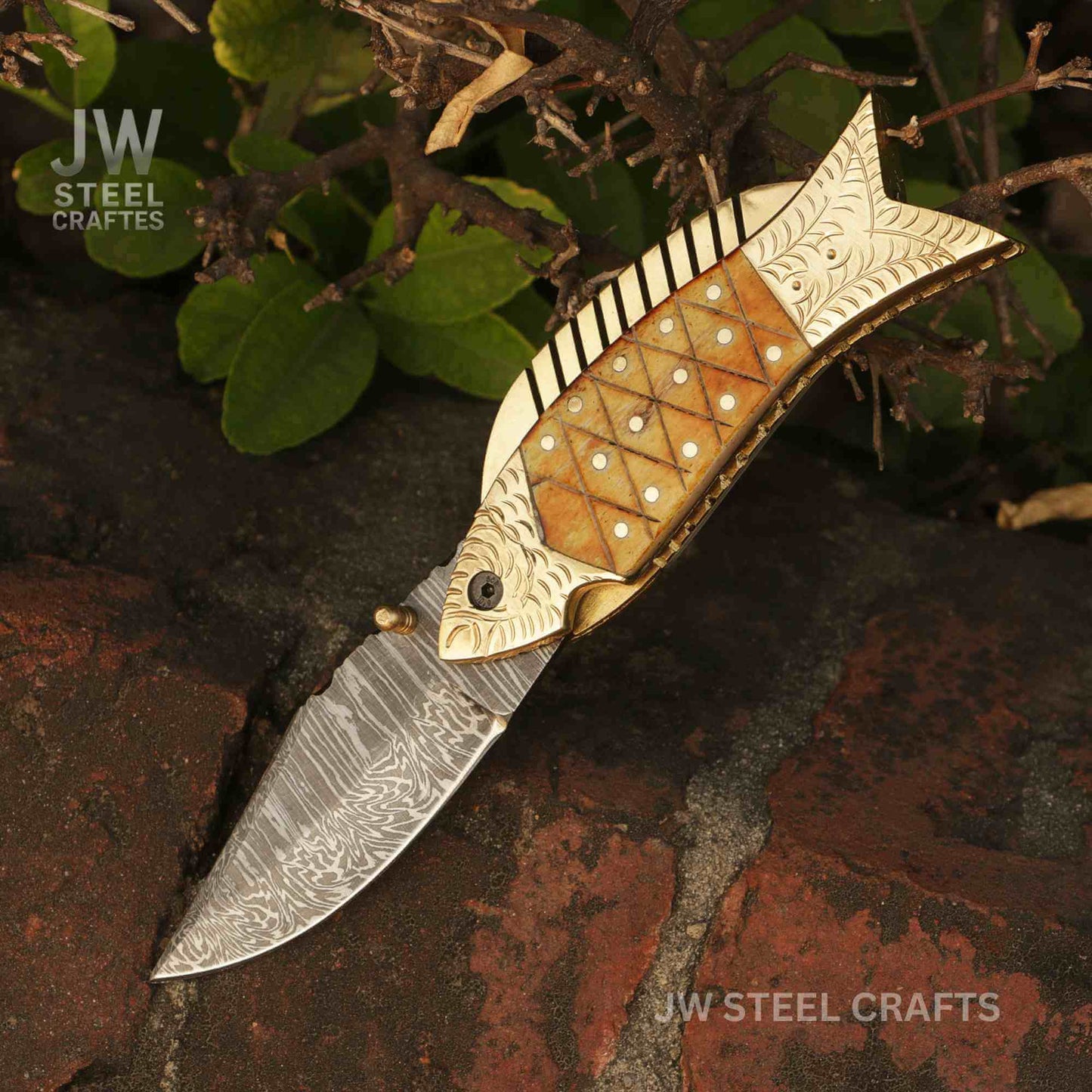 JW SteelCrafts handmade fish-shaped Damascus steel folding knife with engraved brass handle and natural bone inlay, shown open on a stone surface with leaves in a forest setting.