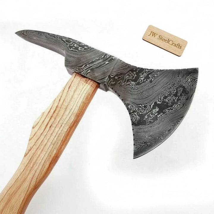     Frostpine custom Damascus survival axe with etched steel blade and smooth pale handle in a woodland campsite environment