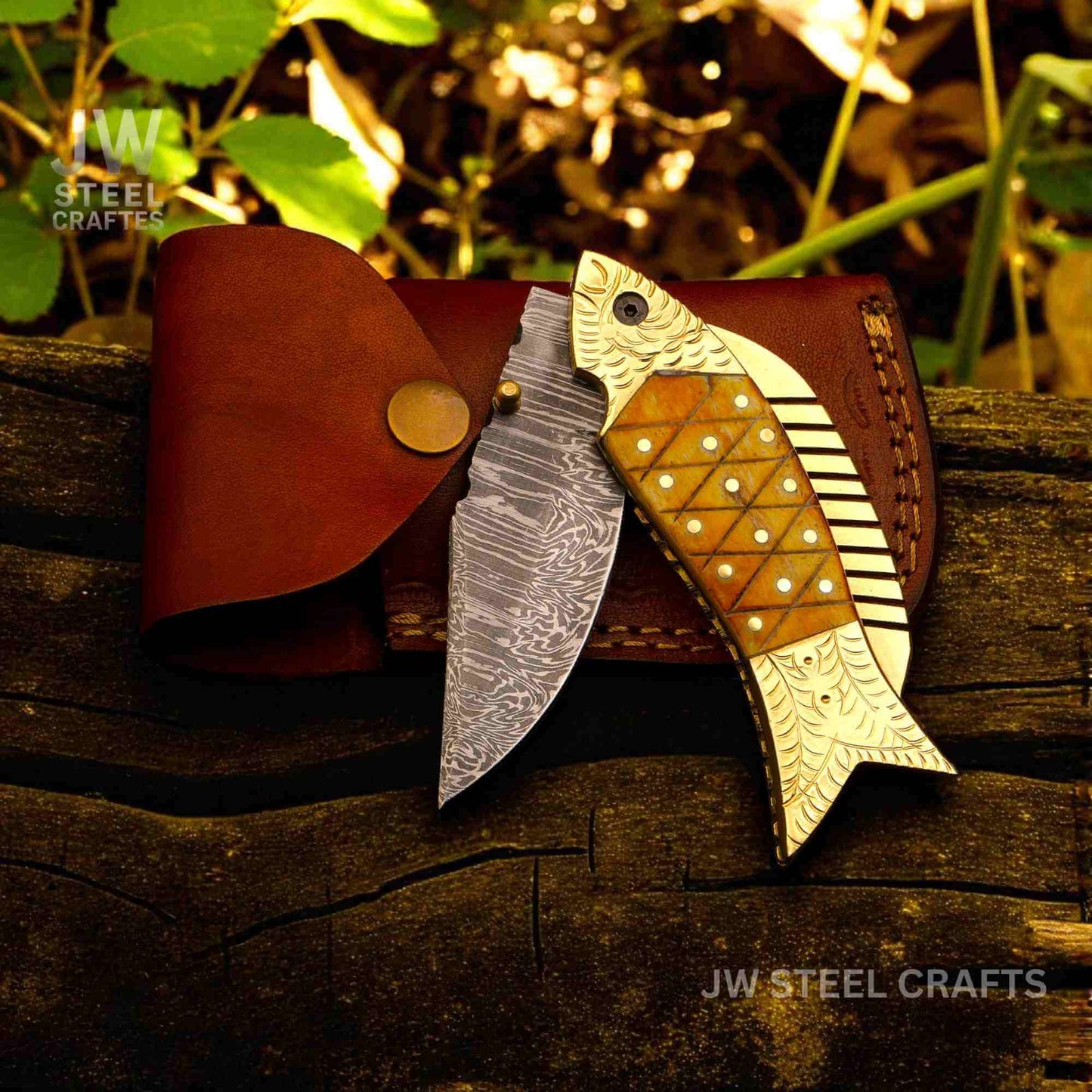 JW SteelCrafts fish-shaped Damascus steel folding knife with engraved brass handle and natural bone inlay, shown open beside a handcrafted brown leather sheath on a wooden log outdoors.