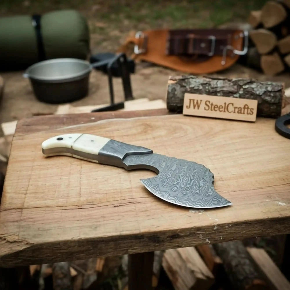     Grayspire custom Damascus curved blade knife with polished ivory handle and detailed steel engravings at a forest campsite.