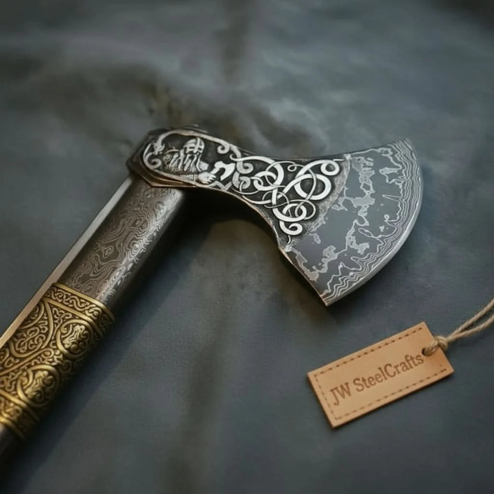     Damascus steel axe and knife set with detailed carvings resting beside weathered logs outdoors.