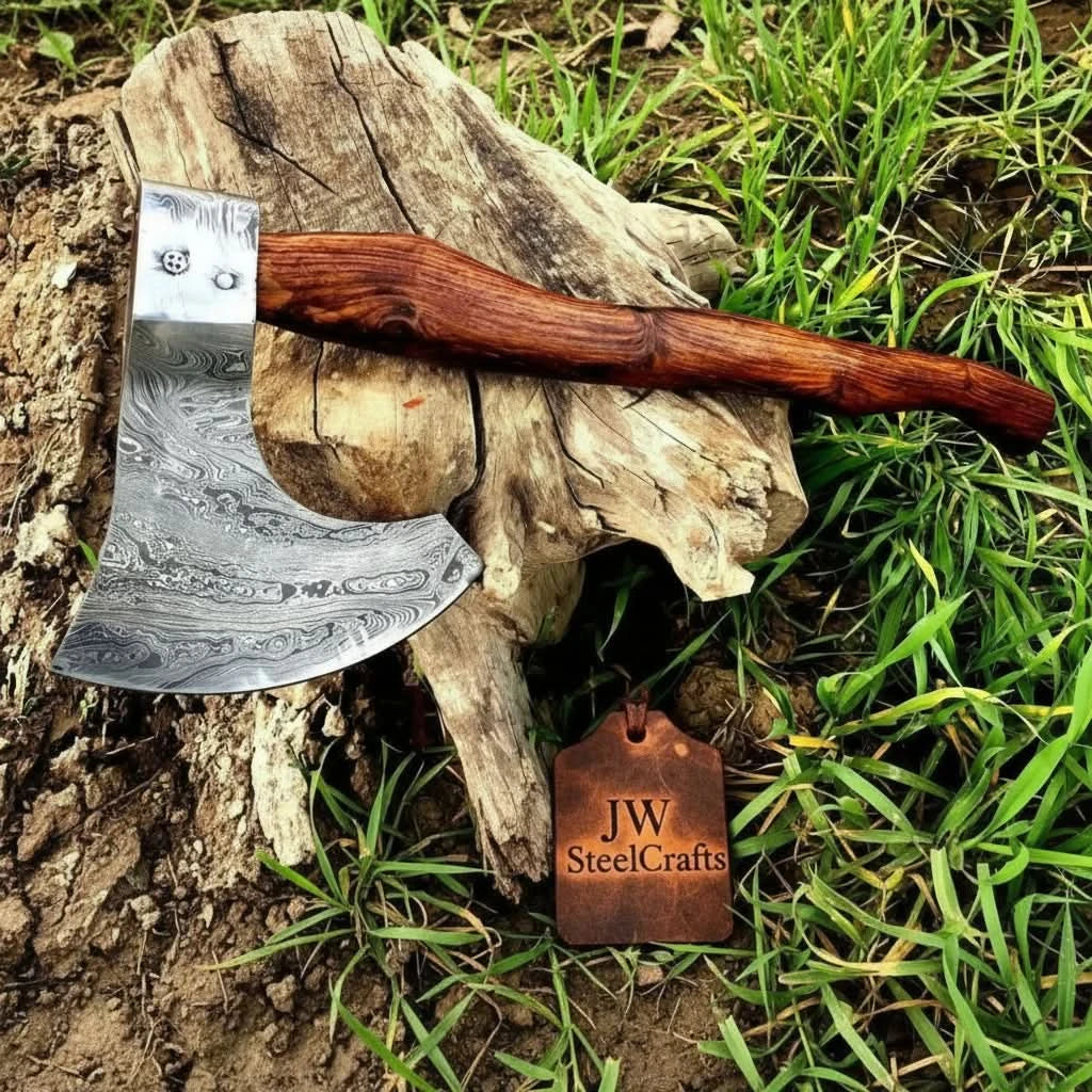     Custom Damascus bushcraft axe featuring engraved steel patterns and a handcrafted hardwood handle displayed naturally on weathered wood in a grassy setting.