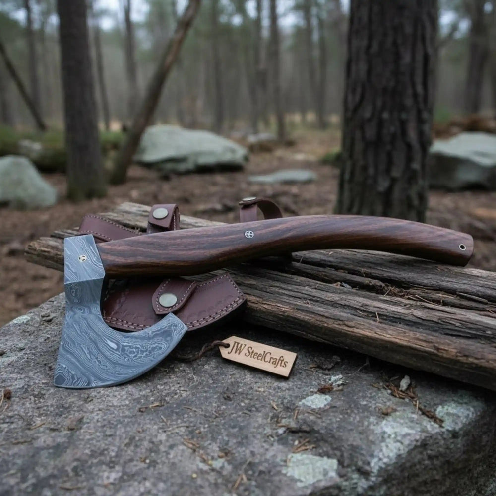     Graniteforge custom bushcraft axe featuring a polished Damascus edge and brown leather sheath in an outdoor forest environment.