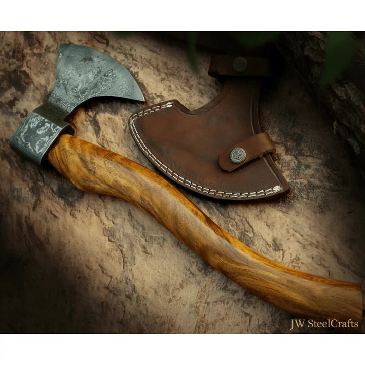     Mistforge Damascus bushcraft hatchet posed on rugged stone and charred timber with elegant handcrafted wooden handle.